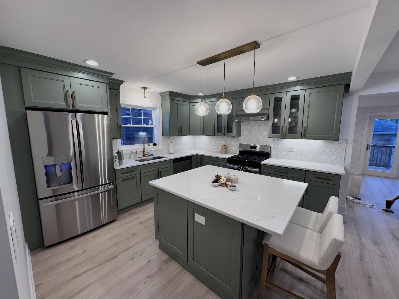 The finished kitchen: sage green shaker cabinets, a white stone island with seating, a marble hexagon backsplash, pendant lights, and light wood floors.